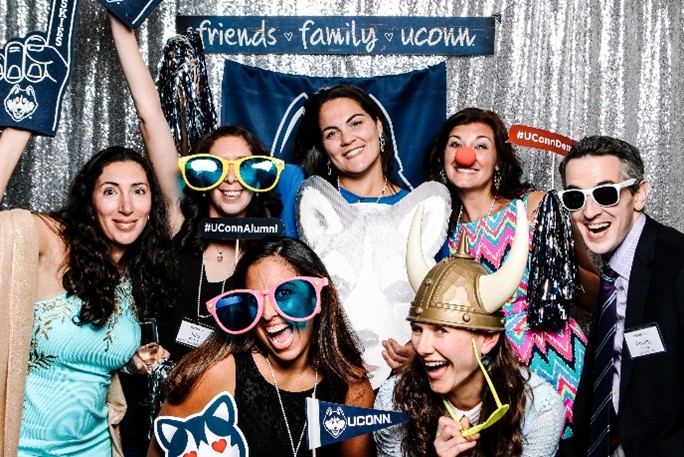 A group of UConn alums at a reunion posing for a photo behind a silver background with a sign that reads "Friends, Family, UConn". They are wearing props such as large glasses, a clown nose, and a Viking hat.