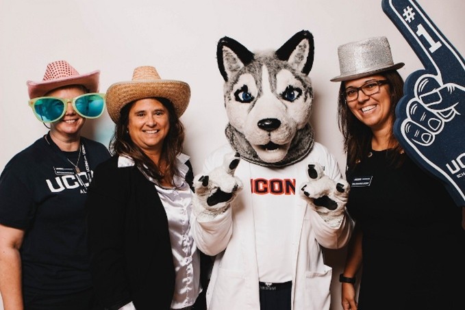 Three people posing with Jonathan mascot in hats and large glasses, with one person holding a navy foam finger.