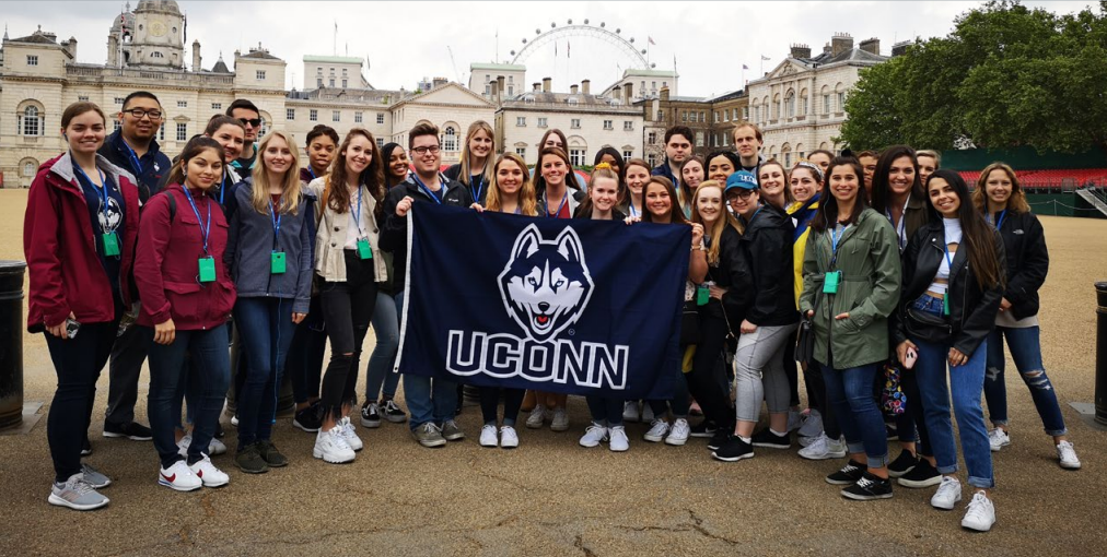 A group shot of UConn graduates posing behind European architecture, holding a UConn flag